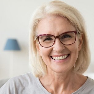 Ältere hellblonde Frau lächelt in die Kamera, dabei trägt sie eine eckige braune Brille